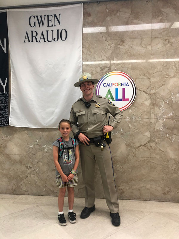 A young girl posing with an officer at the governor's office in front of a sign that reads "California for All"